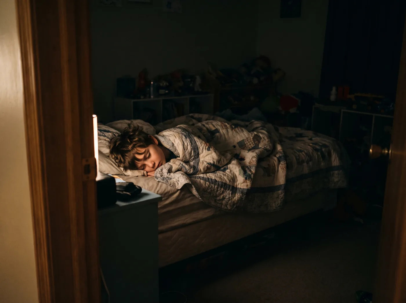 A boy asleep in bed with the blanket pulled up, seen from the doorway of a dark bedroom