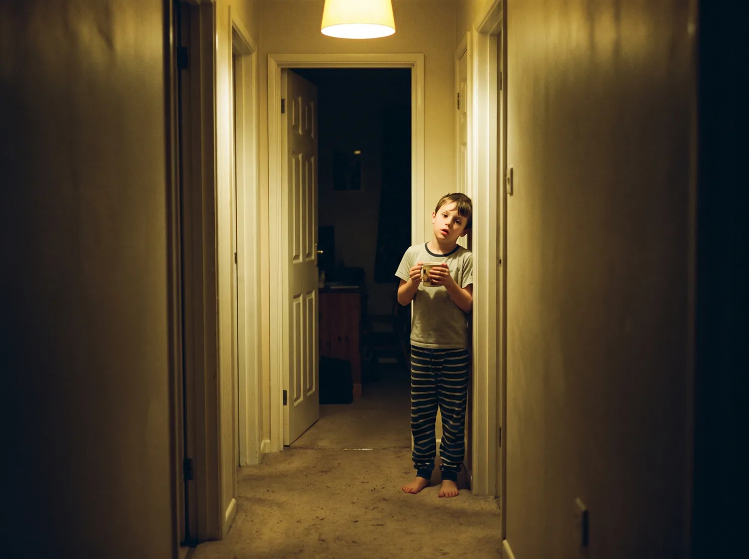 A boy in pajamas standing in a dim hallway at night holding a cup of water, his bedroom door open behind him