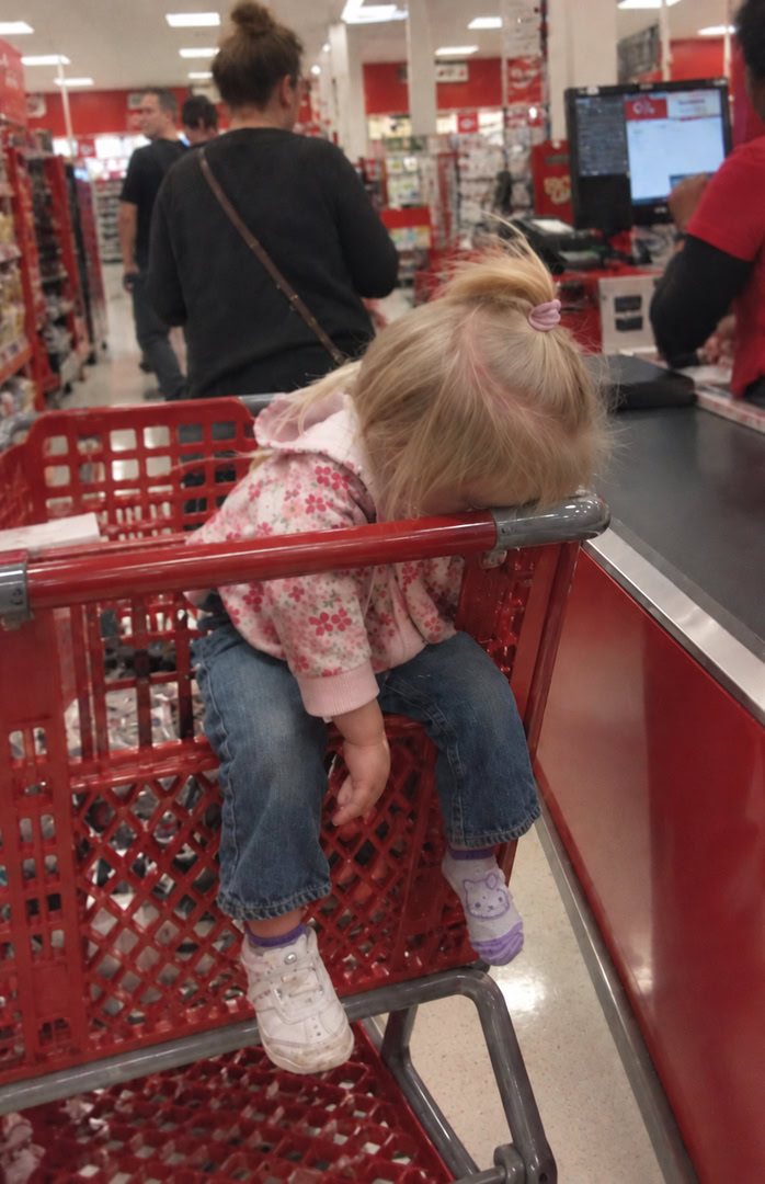 A little girl slumped over the handle of a shopping cart in a checkout line